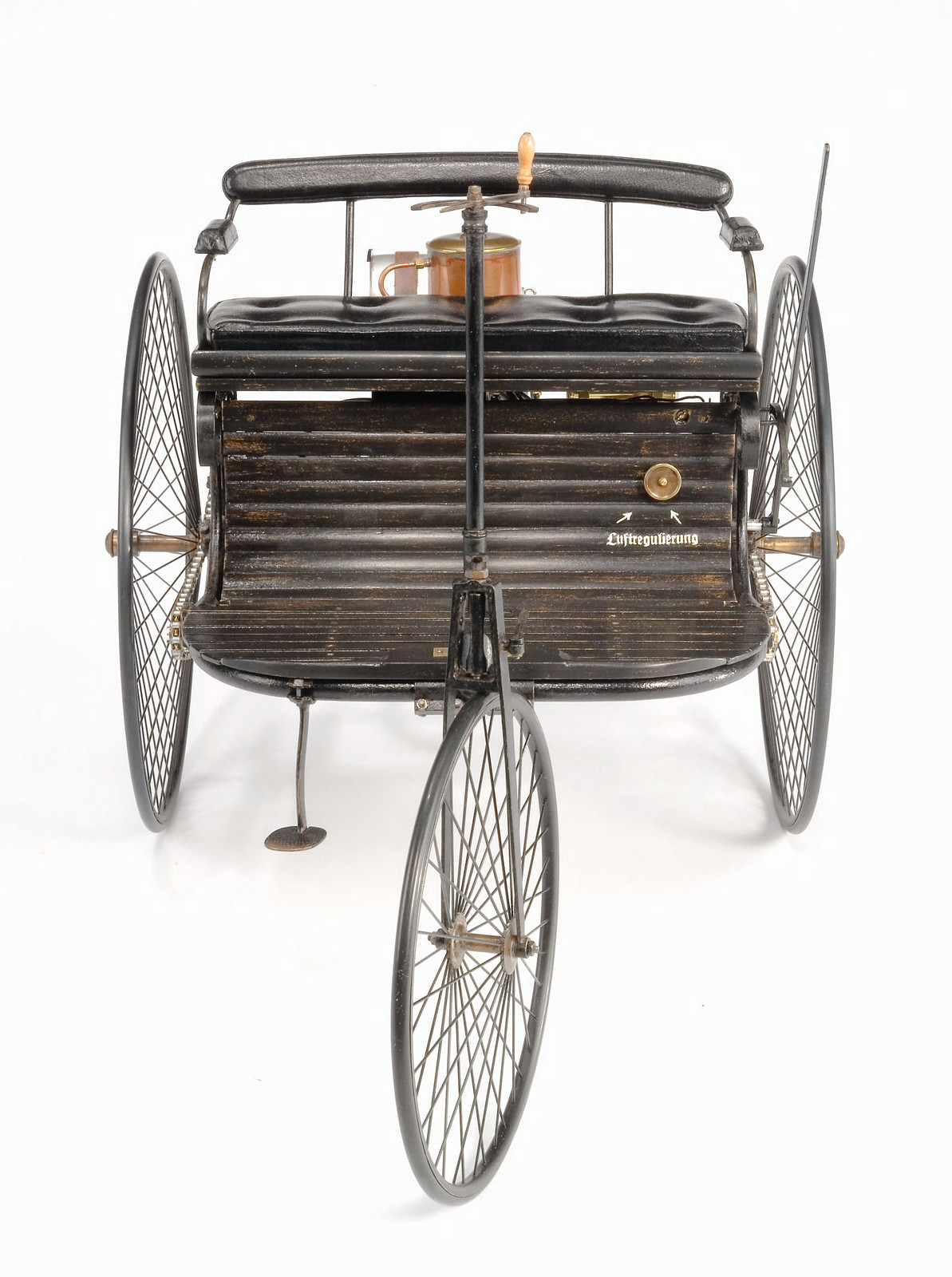 Front view of the preserved three-wheeled Benz Patent-Motorwagen No. 1 artifact against a white background, with its wire wheels, bench seat and tiller visible.