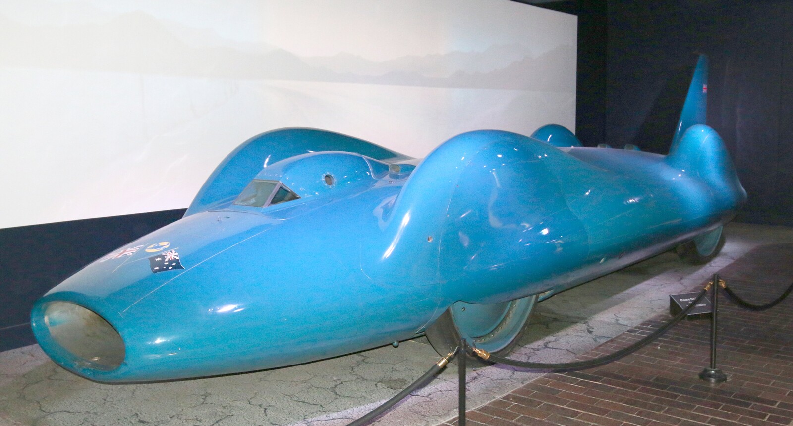 The actual Bluebird CN7 displayed indoors, seen from the front left with its long blue body, enclosed wheels, cockpit and tall stabilizing tail visible.