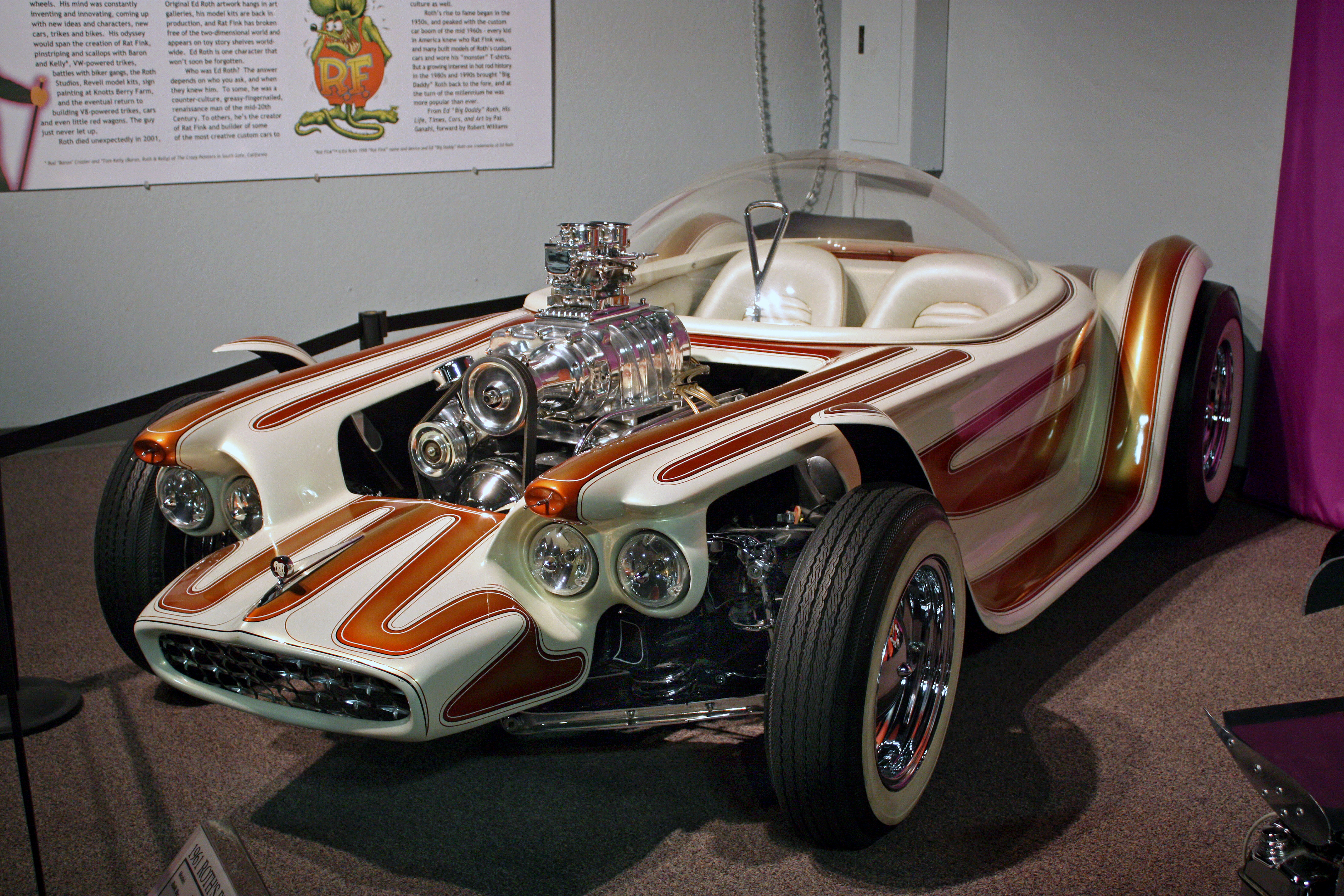 The actual restored 1961 Beatnik Bandit displayed indoors, with its transparent bubble canopy, white-and-gold body and exposed supercharged engine visible.