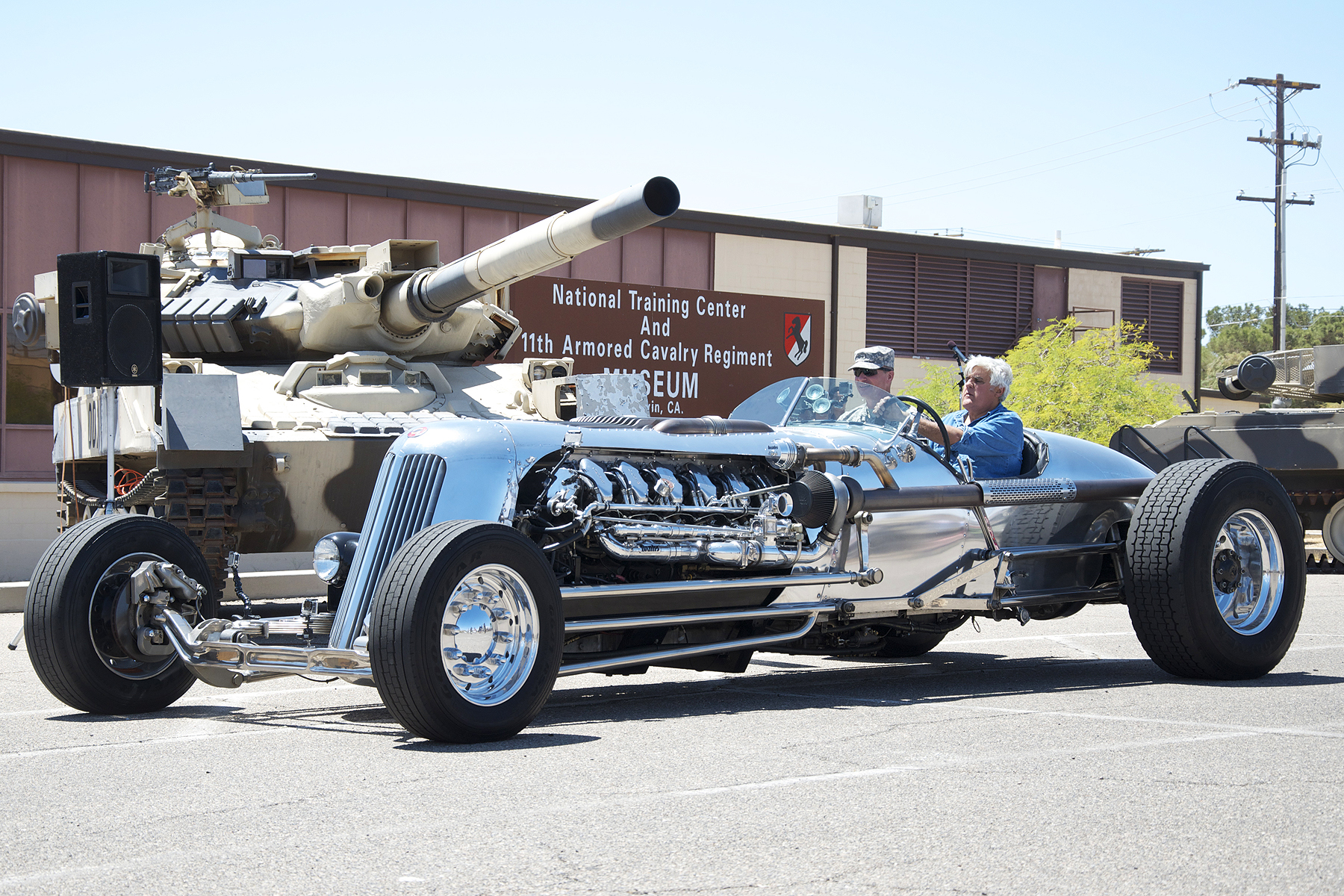 Jay Leno driving the polished aluminum Blastolene Special at Fort Irwin, with its exposed V12 and a military tank visible behind it.