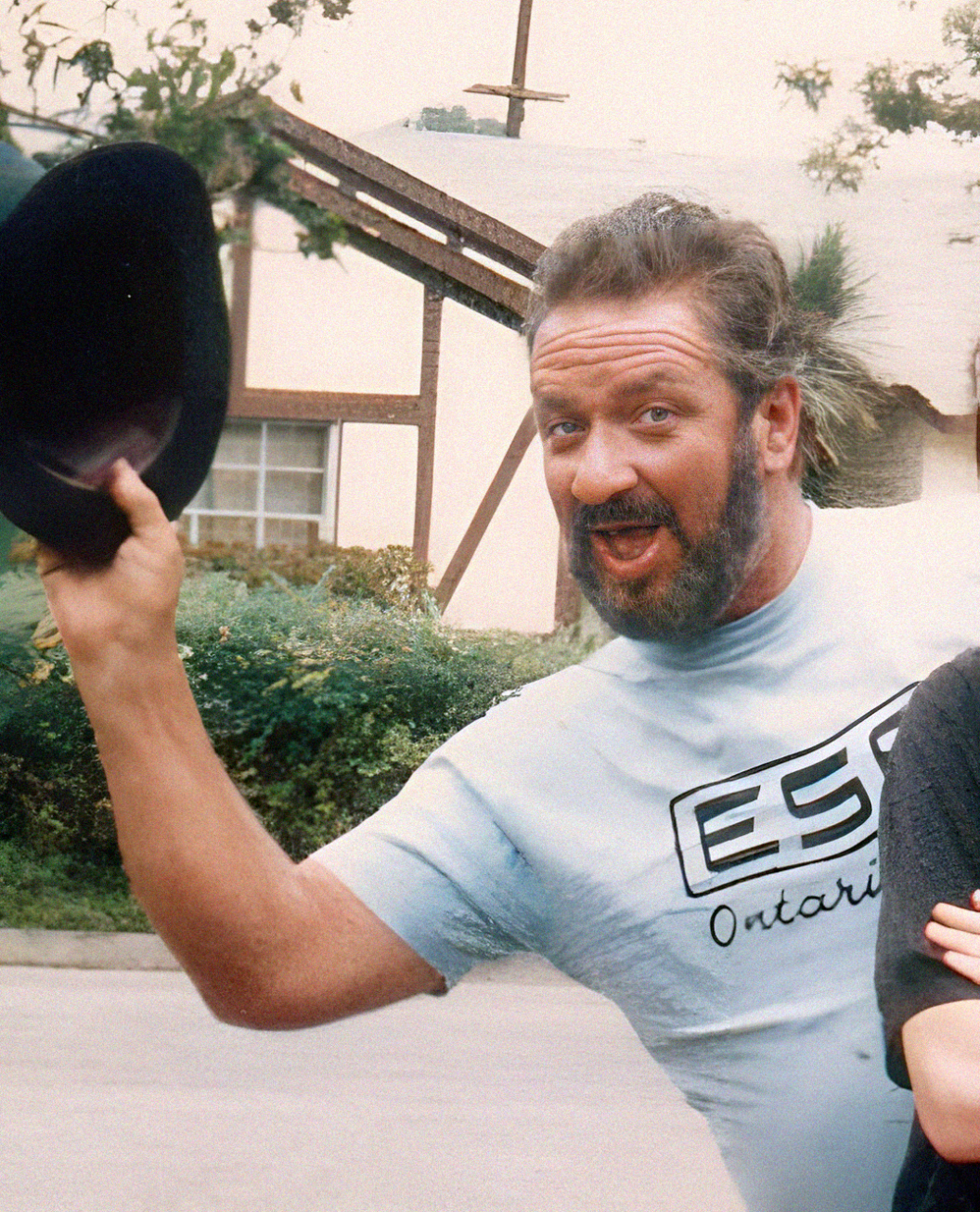 Ed “Big Daddy” Roth in a 1987 outdoor portrait, lifting a black hat and wearing a light-blue T-shirt.