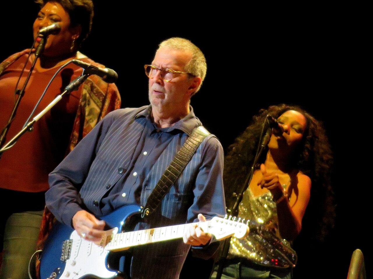 Eric Clapton playing electric guitar onstage in London in 2018, with two backing vocalists behind him.
