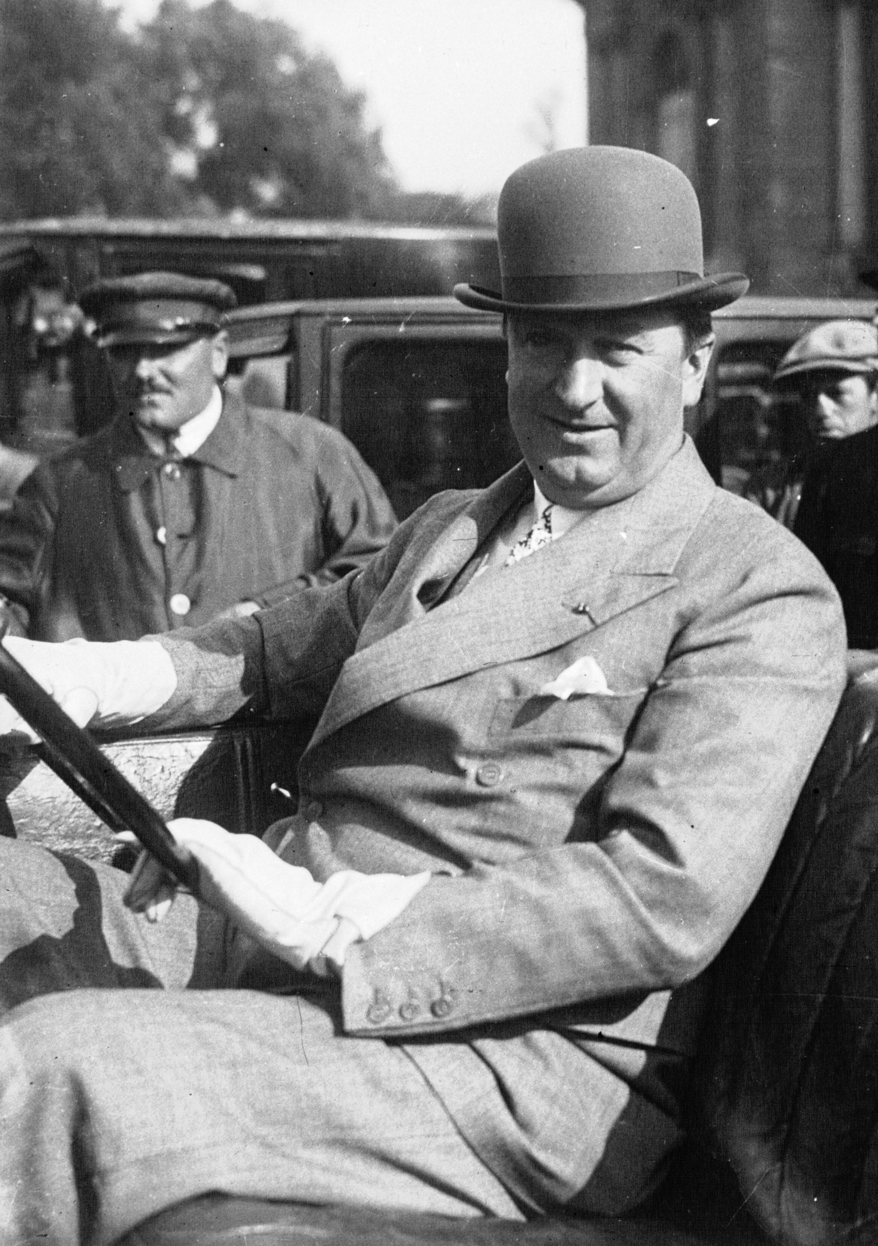 Black-and-white 1926 photograph of automobile designer Ettore Bugatti seated at the wheel wearing a bowler hat and suit.