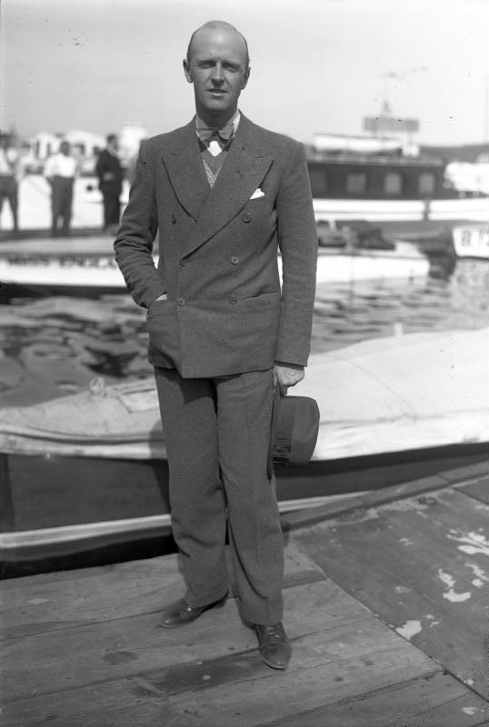 Black-and-white full-length portrait of Sir Henry Segrave in a suit standing beside a motorboat.
